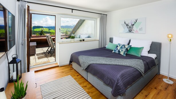 A bedroom with a bed, a window with a view, a potted plant, and a floor lamp. - Oberreute - Modern vacation apartment with panoramic views of the Allgäu (Oberreute)