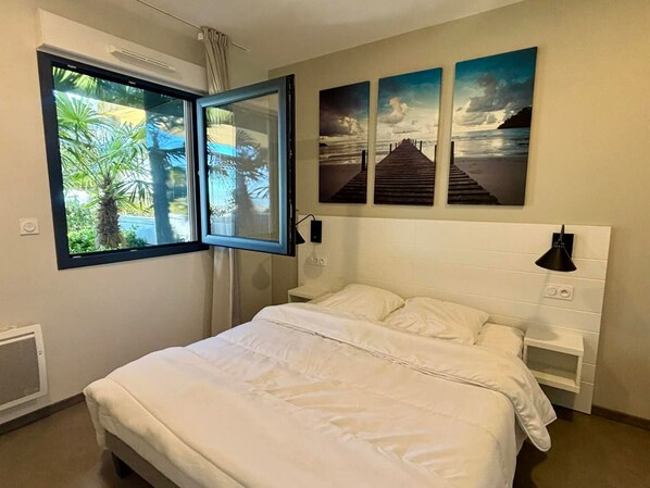 Quarto com cama, janela com vista para palmeiras e três quadros na parede.