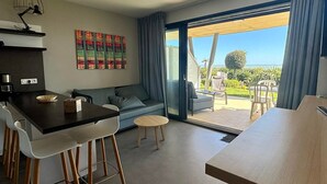 Un salon moderne comprenant un canapé, une table à manger et des chaises, un coin cuisine avec une machine à café, et une grande porte-fenêtre coulissante donnant sur une terrasse extérieure avec vue sur l’océan.