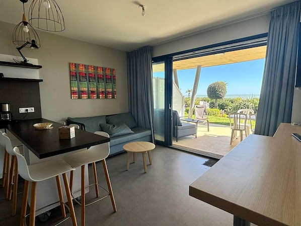 A modern living room with a sofa, dining table, and chairs, a kitchen area with a coffee machine, and a large sliding glass door leading to an outdoor patio with a view of the ocean.