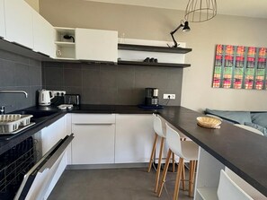 Una cocina moderna con armarios blancos, encimera negra, una cafetera y un comedor con sofá azul y sillas blancas.