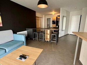 Uma sala de estar moderna com um sofá azul, uma mesa de centro de madeira e uma área de cozinha com um banquinho e micro-ondas.