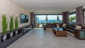 Ein modernes Wohnzimmer mit einem großen Flachbildfernseher, einer dunkelbraunen Sofagarnitur, einem Glastisch und Meerblick durch große Fenster.