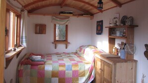 A cozy bedroom with a wooden ceiling, a bed with a colorful quilt, a small window with a view, and a shelf with kitchen items. - Cosy Woodland off Grid Shepherds Hut - Hazel (Castle Douglas)