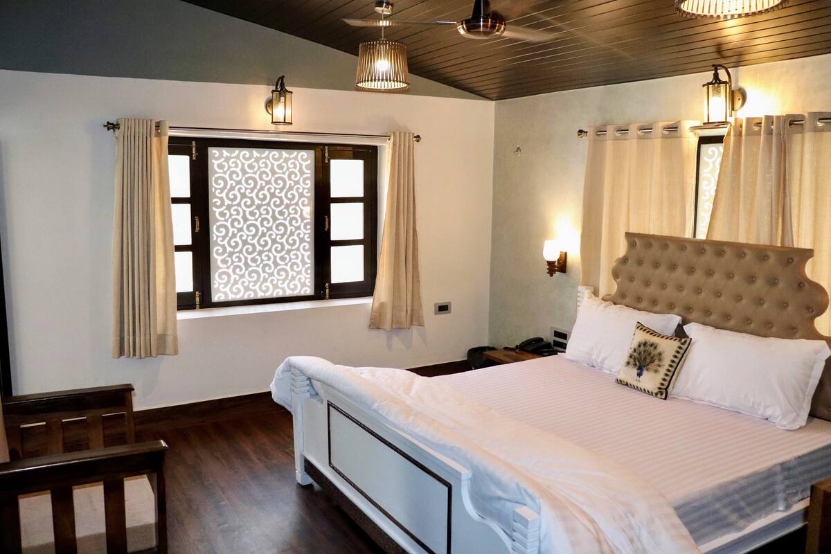 A bedroom with a large bed, wooden headboard, and a window with curtains. There is a nightstand with a lamp and a telephone.