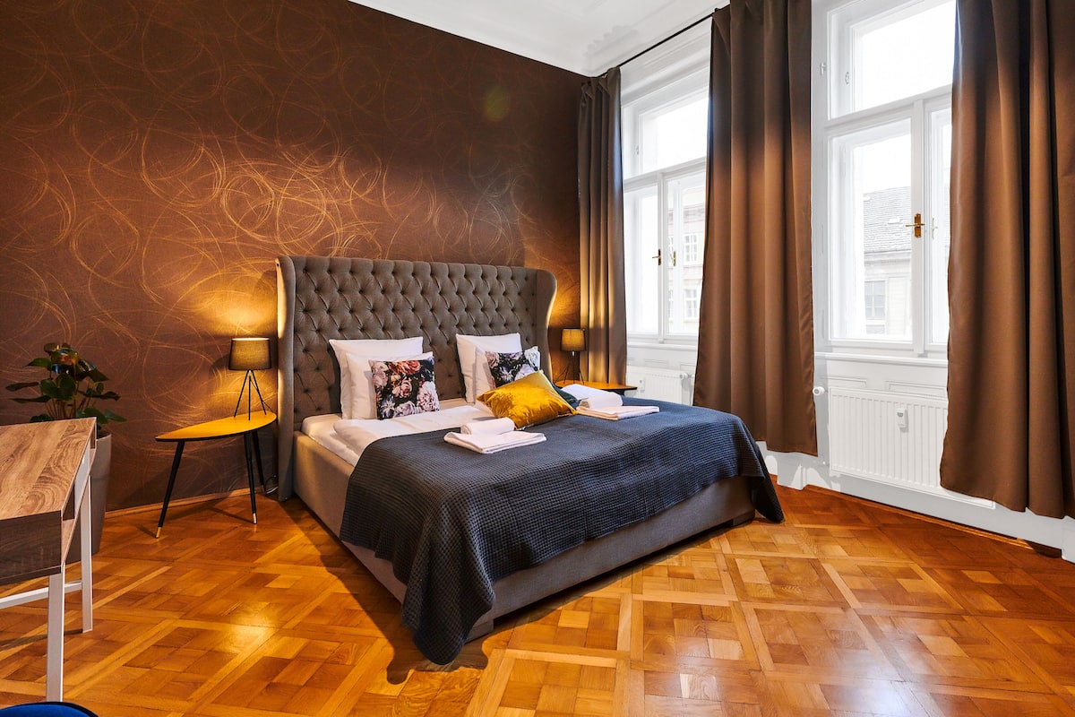 Ein Schlafzimmer mit einem gepolsterten Kopfteil, Holzboden, einem Bett mit dunkelblauer Bettdecke und einem Fenster mit Vorhängen.