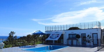 A pool area with a lounge building, a covered seating area, and a view of the ocean.