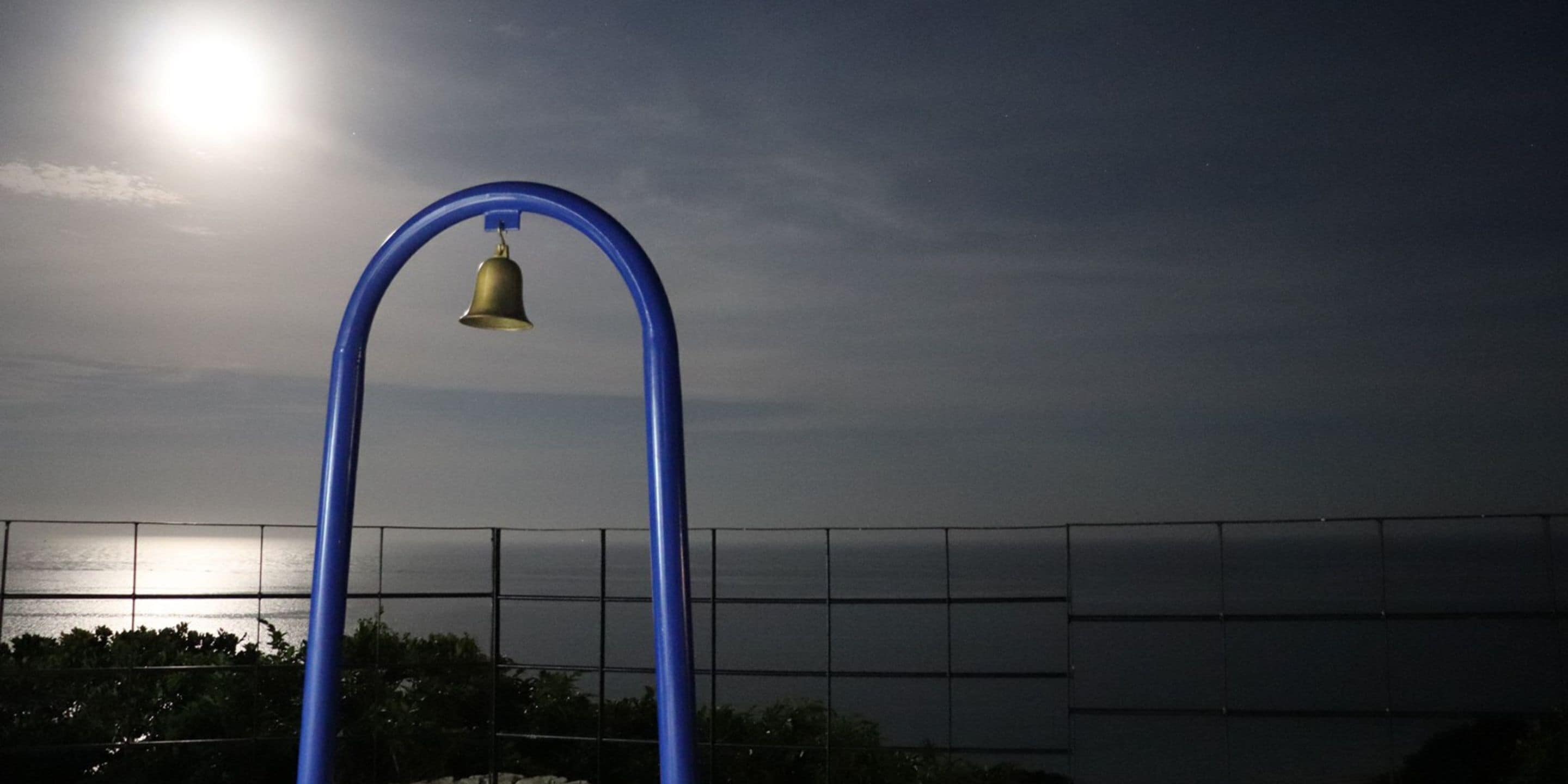 Eine blaue Metallkonstruktion mit einer Glocke, vor einem Sonnenuntergang über dem Wasser, mit einem Zaun im Vordergrund.