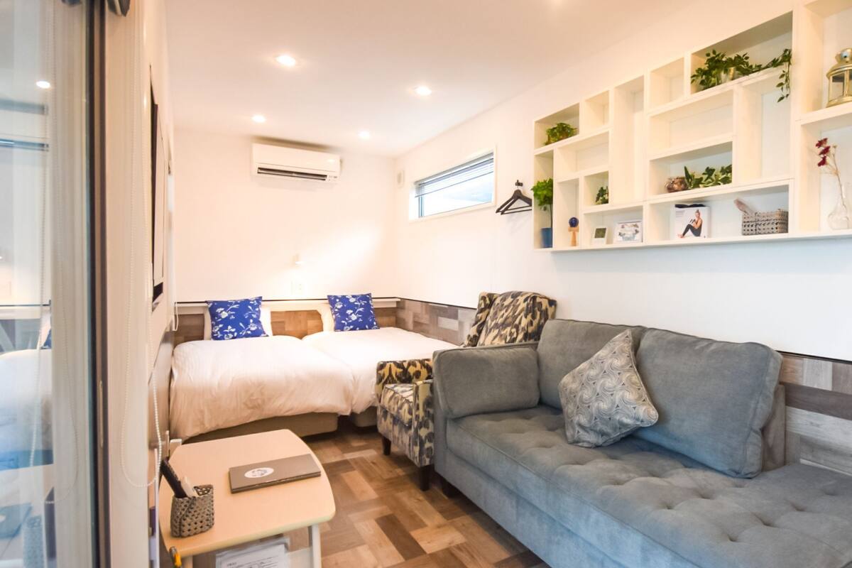Une chambre d’hôtel compacte comprenant deux lits, un canapé, une petite table et une étagère murale agrémentée de plantes et d’objets décoratifs.