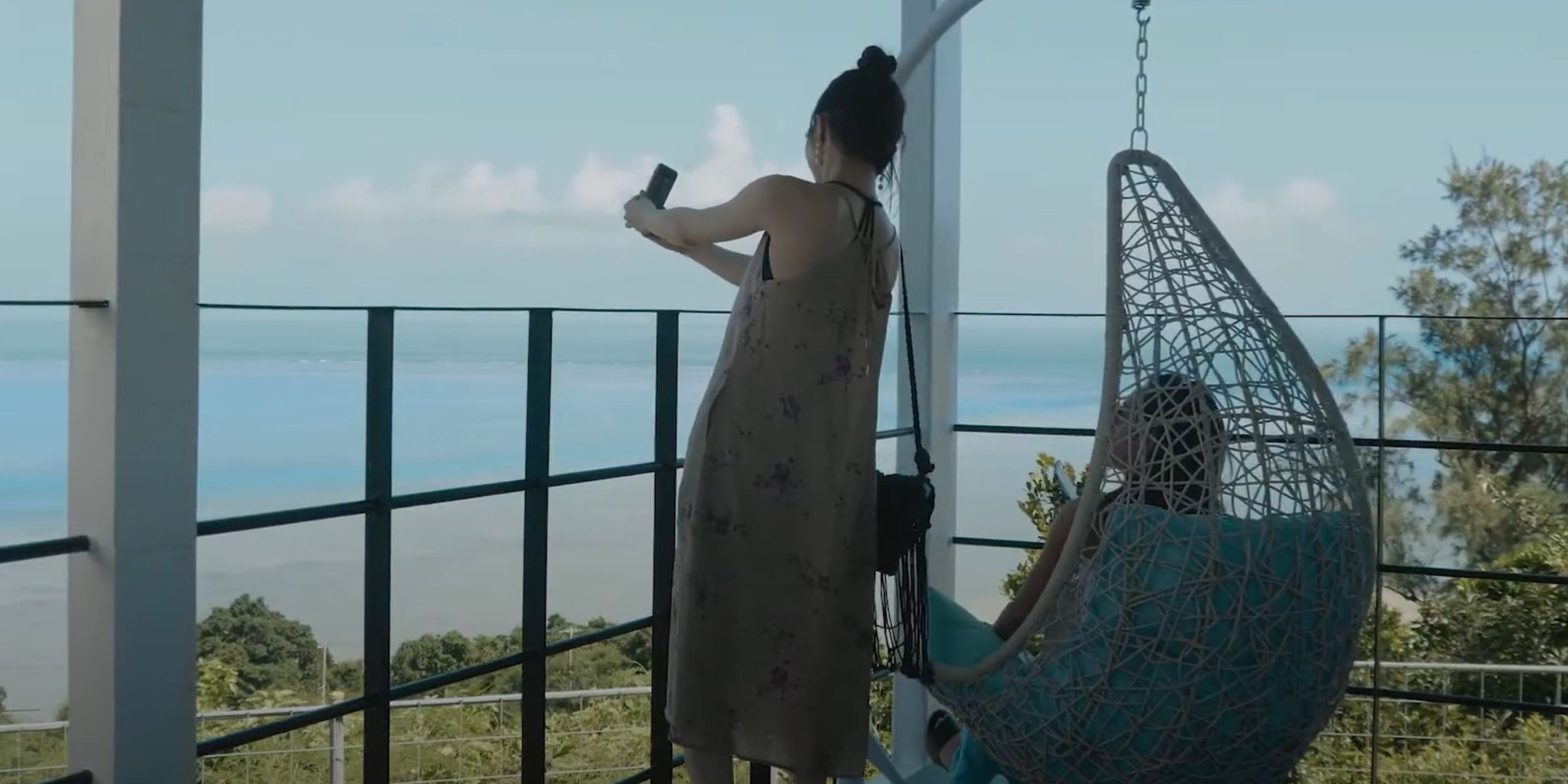 Una persona tomando una foto con un teléfono inteligente, otra persona sentada en una hamaca colgante y vista al mar.