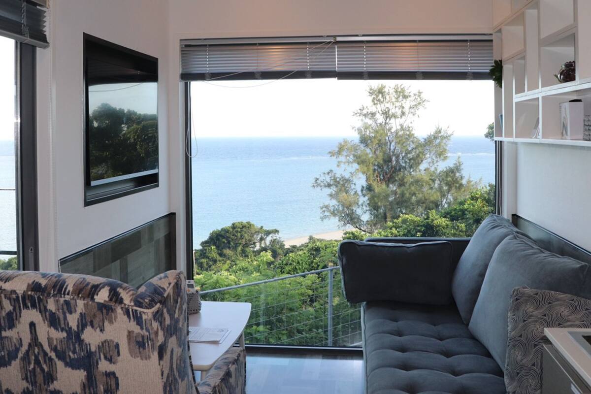 Un salon moderne avec un canapé, un fauteuil à motifs et un téléviseur. Il est doté d’une grande fenêtre offrant une vue sur la mer et la verdure.