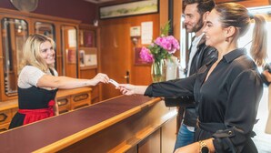 A reception area with a wooden counter, a clock on the wall, and a vase with flowers. - Apartmenthaus Hotel Cella Central (Zell am See)
