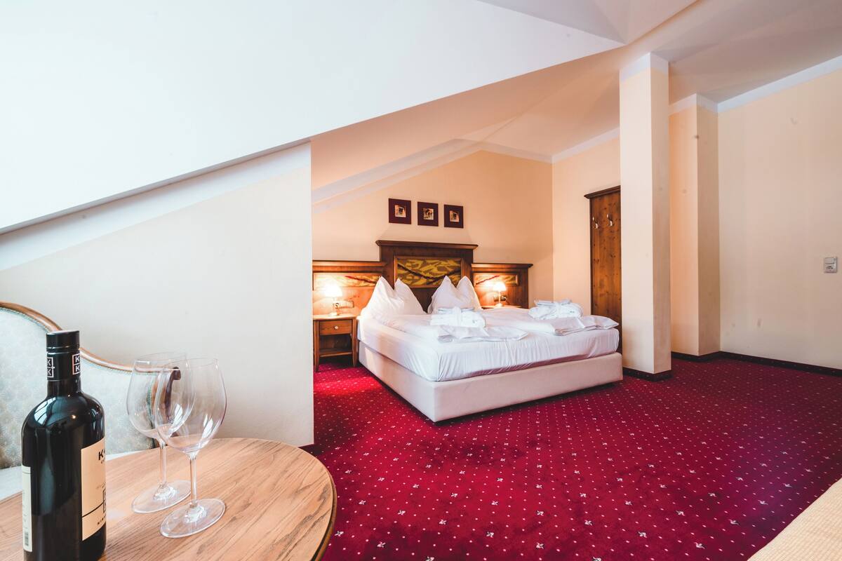 Habitación de hotel con alfombra roja, cama con ropa de cama blanca, mesita de noche con una botella y vasos, y cabecero de madera.