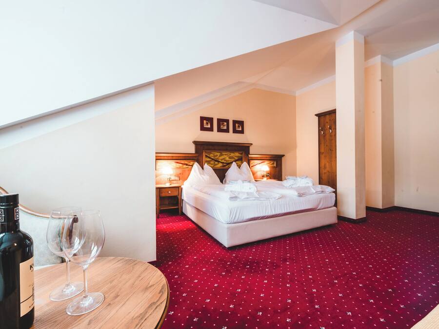 Habitación de hotel con alfombra roja, cama con ropa de cama blanca, mesita de noche con una botella y vasos, y cabecero de madera.
