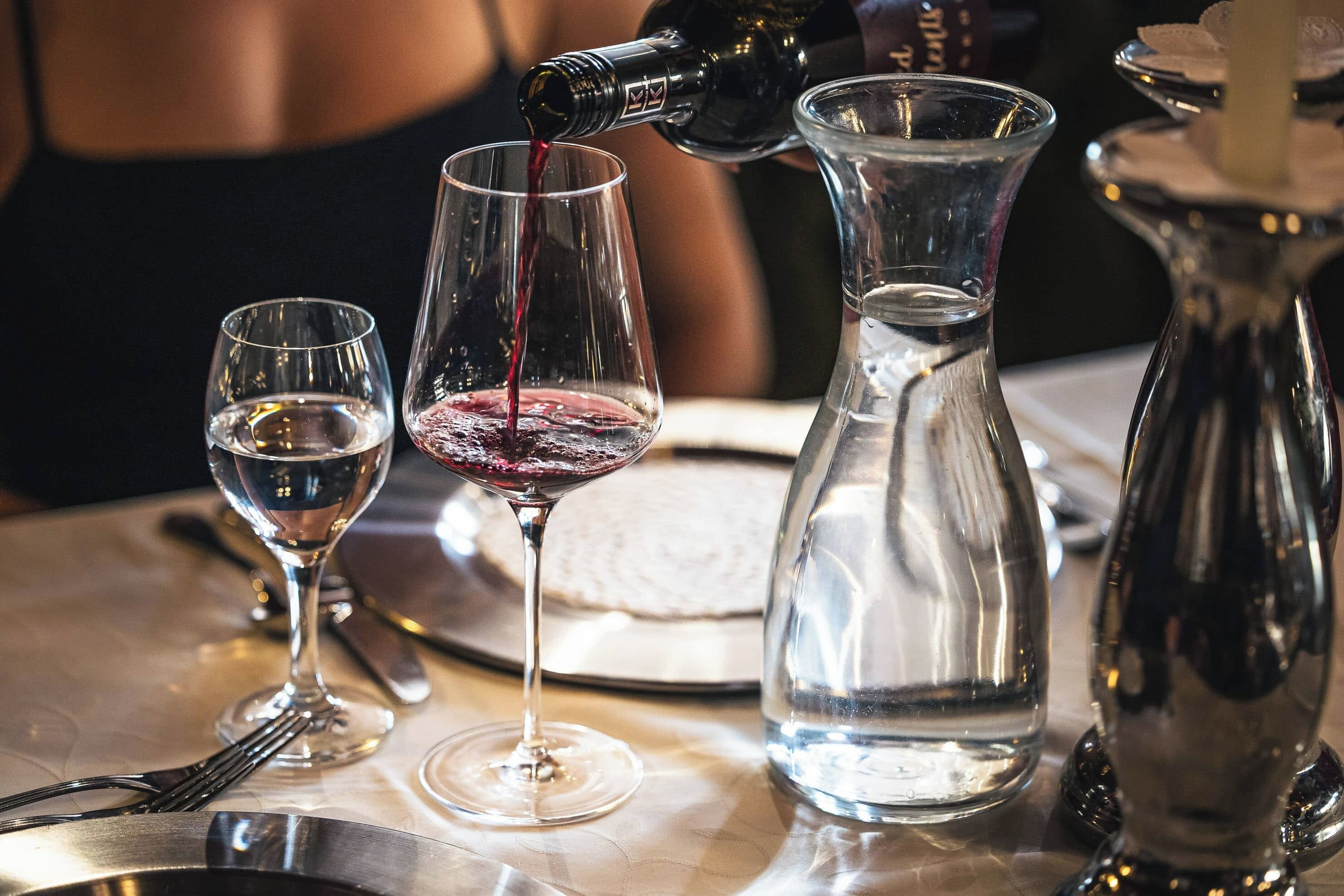 Uma mesa com copos de vinho, jarra de água e uma garrafa de vinho sendo servida.