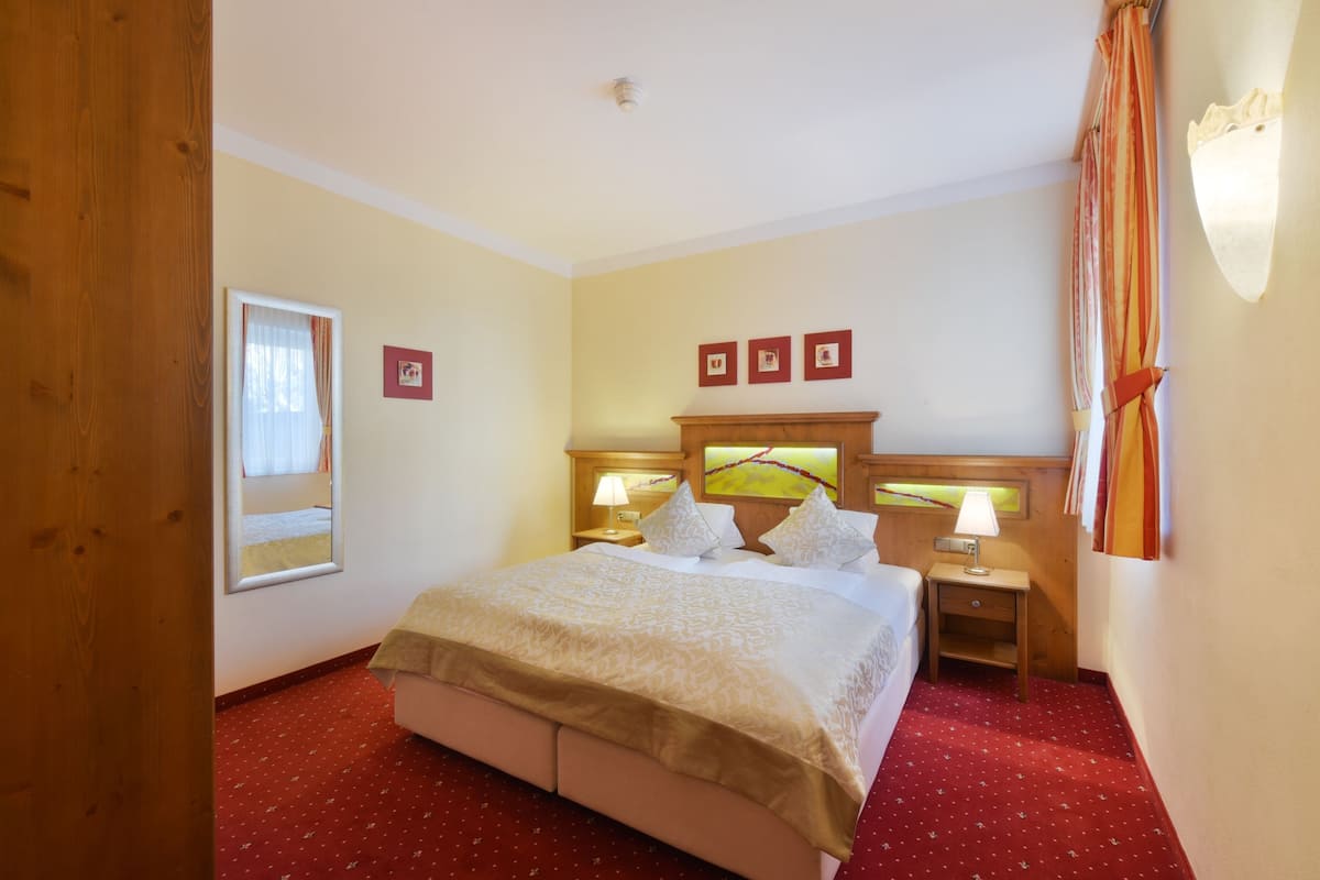 Habitación de hotel con cama, dos mesitas de noche con lámparas, cabecero de madera y alfombra roja.