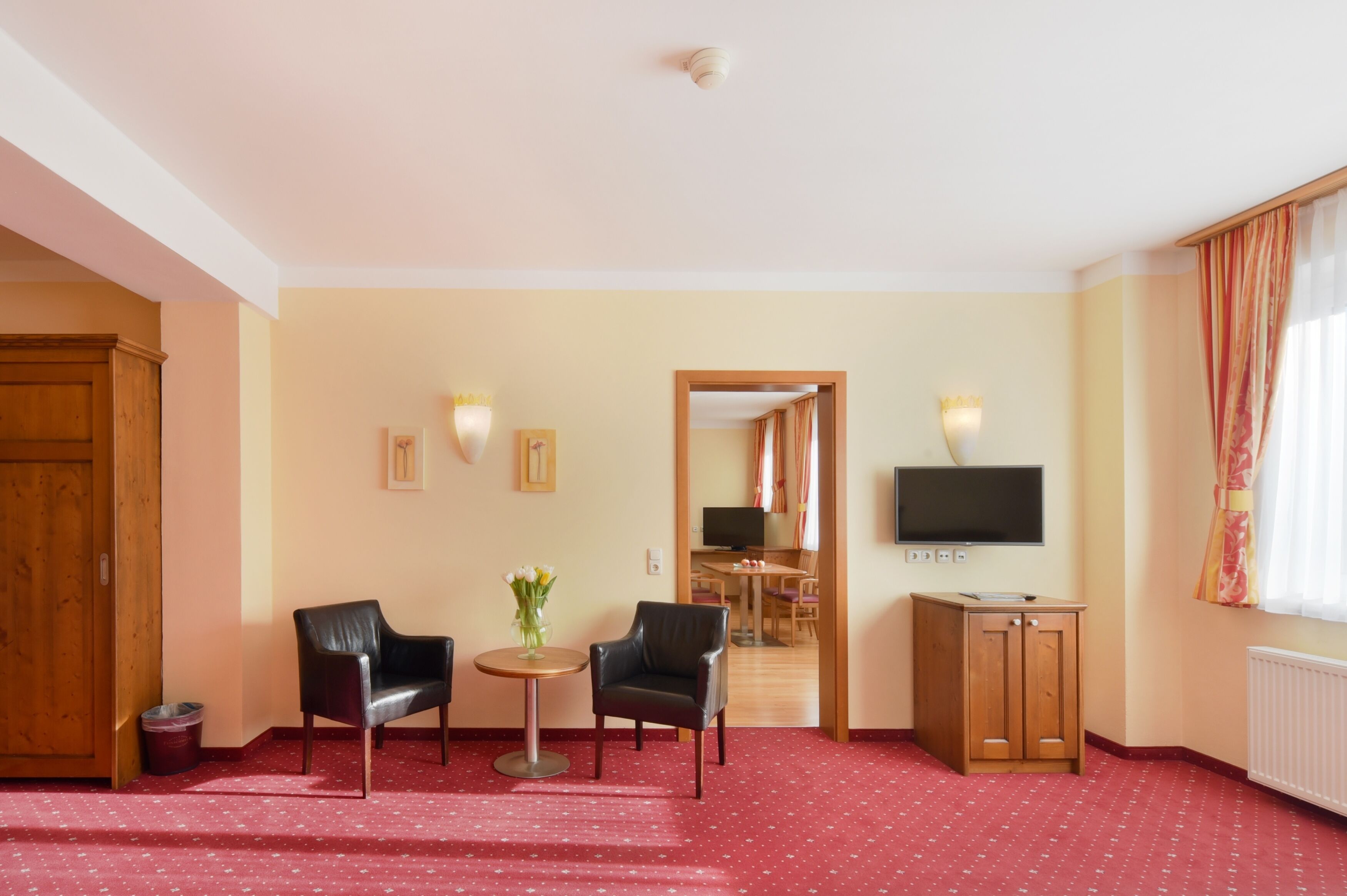 Quarto de hotel com carpete vermelho, duas poltronas de couro preto, uma mesinha redonda com um vaso de flores, um armário de madeira, uma televisão fixada na parede e uma porta de madeira.