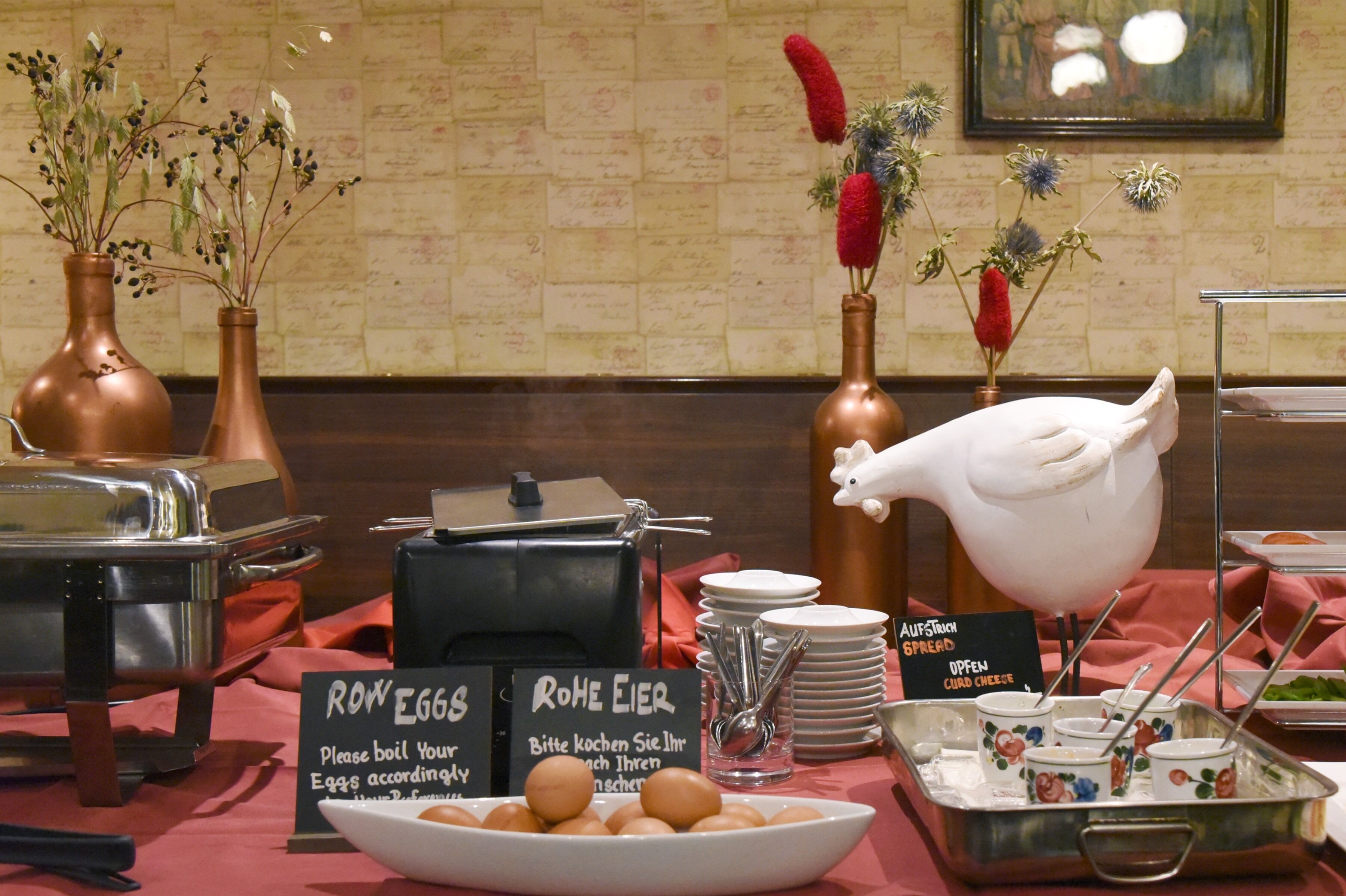 Un buffet de petit-déjeuner avec des plats étiquetés, notamment des œufs et du fromage, ainsi que des vases décoratifs remplis de fleurs.
