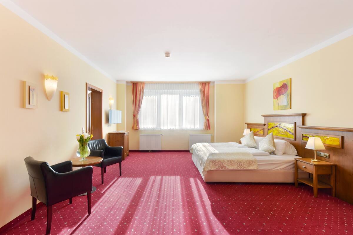 Una habitación de hotel con cama, escritorio, silla, lámpara y ventana con cortinas.