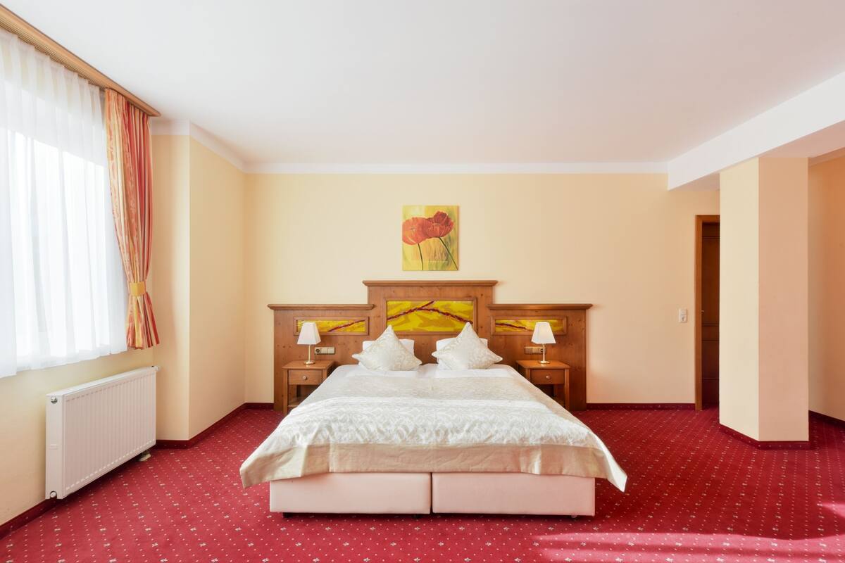 Habitación de hotel con una cama grande, dos mesitas de noche con lámparas, un radiador y una ventana con cortinas.