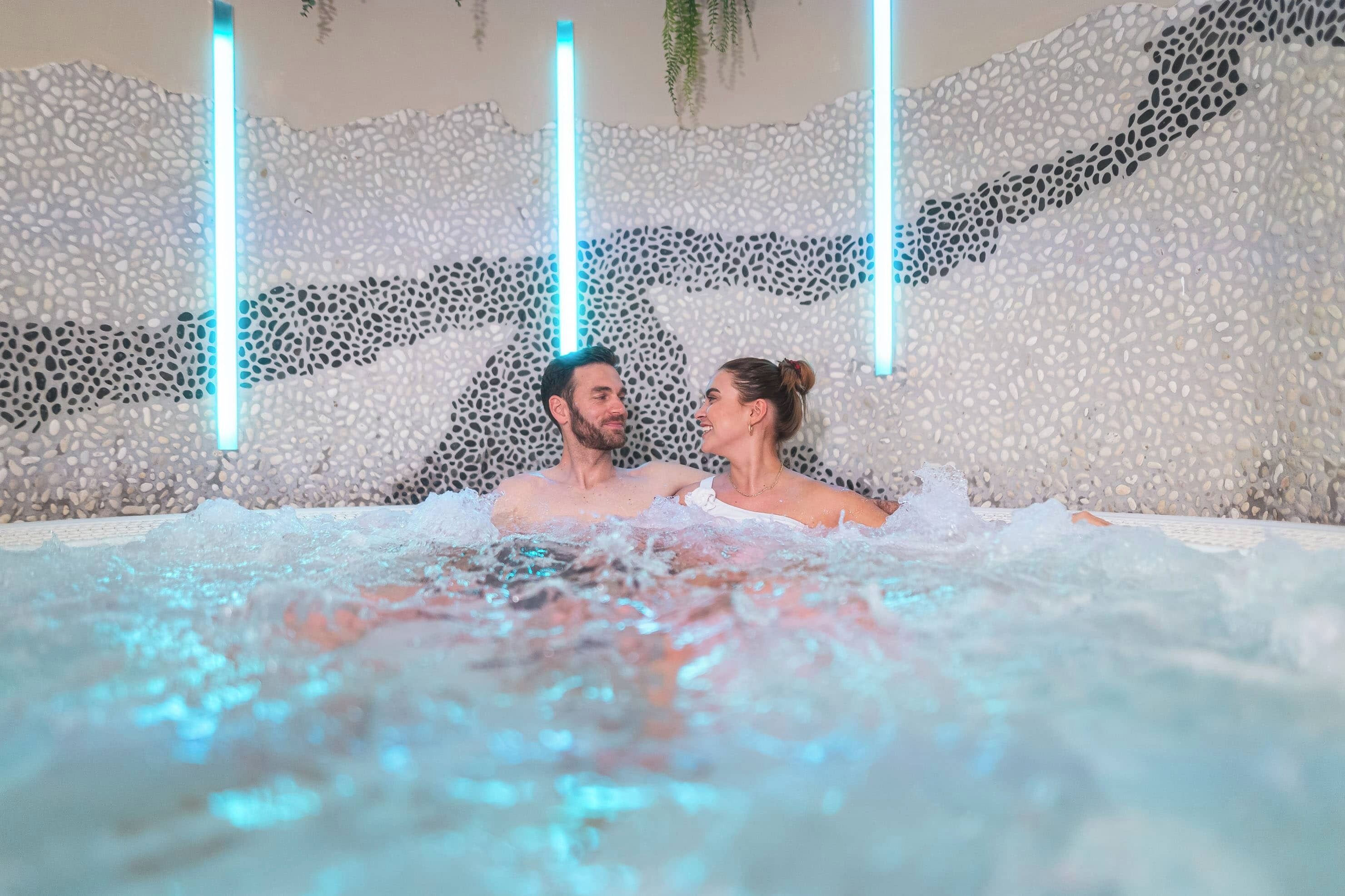 Una pareja en un jacuzzi con pared de piedras y luces verticales azules.