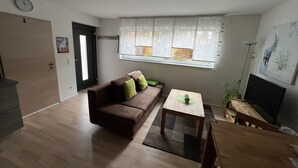 A living room with a brown sofa, a small table, a TV, and a window with patterned curtains. - High Franconia apartment  (Oberkotzau)