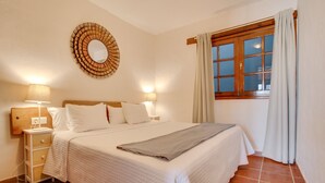 Ein ordentlich bezogenes Bett mit Kopfteil, zwei Nachttischlampen, einem Nachttisch, einem Spiegel und einem Fenster mit Vorhängen.