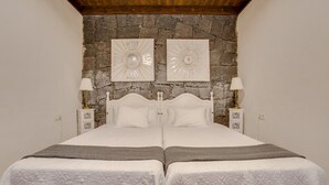 Uma cama de casal com parede de pedra ao fundo, duas mesas de cabeceira com abajures e teto com vigas aparentes.