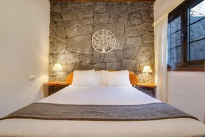 Un dormitorio con pared de piedra, una cama con ropa de cama blanca, dos mesitas de noche con lámparas y una ventana con cortinas.