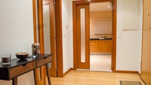 A hallway with a wooden floor, a black console table with a glass, a vase, and a bowl. A wooden door leads to a kitchen area with wooden cabinets and a countertop. - JOIVY Graca Luminous 2-BR Apartment w/Terrace (Lisboa)