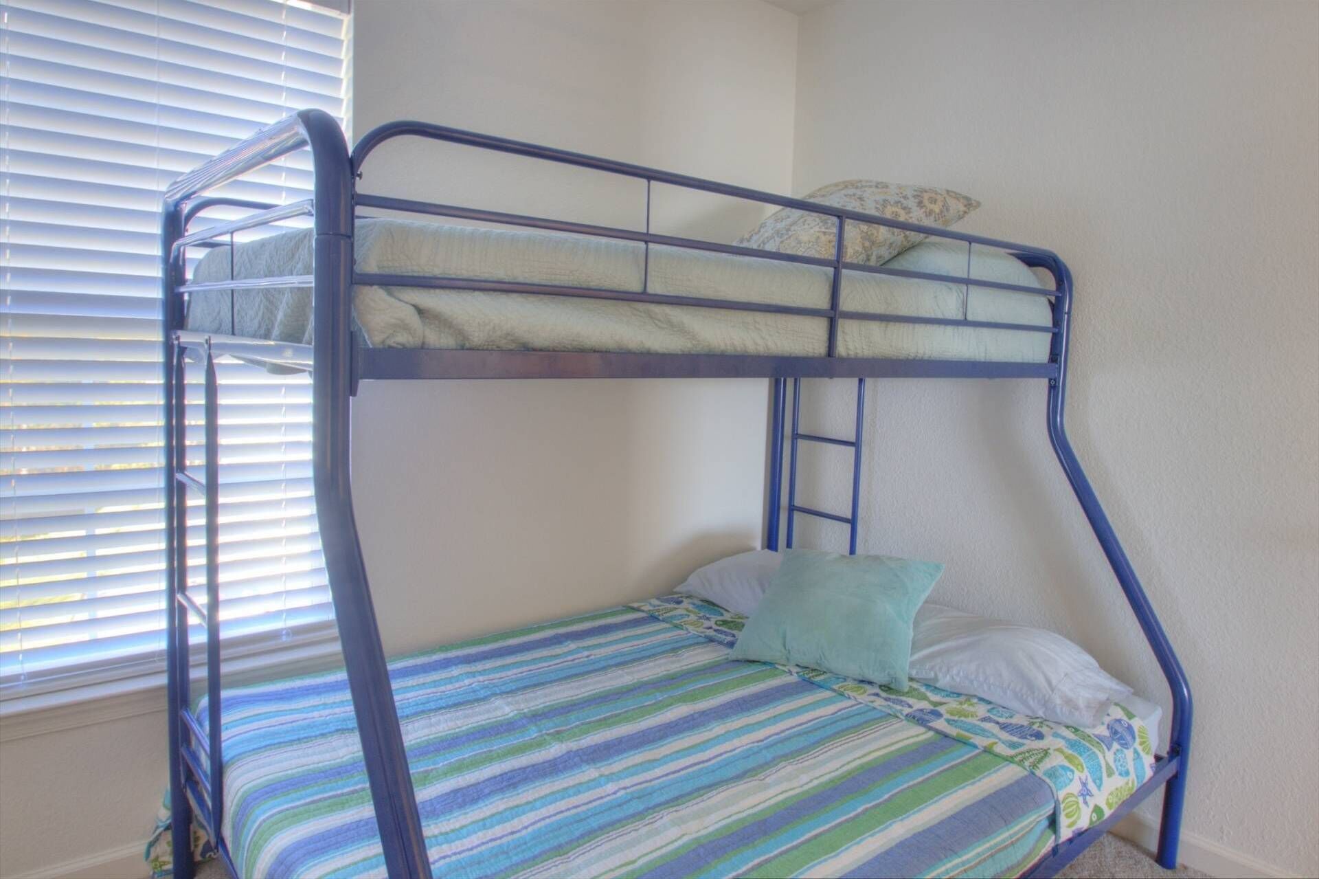 A bunk bed with a striped bedspread and pillows, a ladder, and a window with blinds.