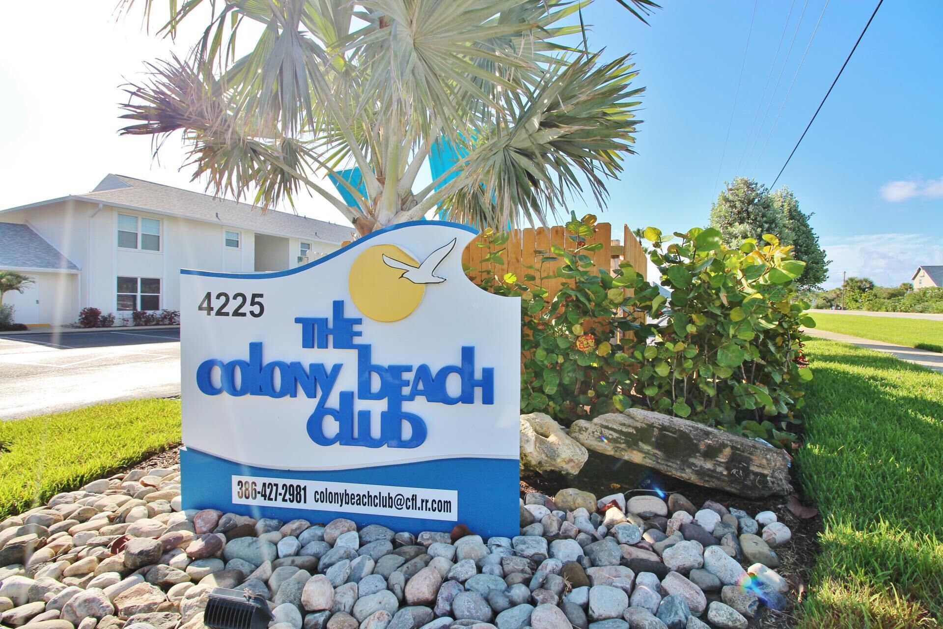 A sign for The Colony Beach Club with a beachside view, including a house and palm trees.