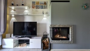 A living room with a flat-screen TV, a fireplace, and a shelf with decorative items. - Lilja Vacation Home: Large Garden, Fireplace, Stone Grill, Dog-Friendly, Outdoor Pool, Beach Package (Trzęsacz)