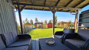 An outdoor patio with wicker furniture and a table, overlooking a well-maintained lawn and a wooden structure in the background. - Lilja Vacation Home: Large Garden, Fireplace, Stone Grill, Dog-Friendly, Outdoor Pool, Beach Package (Trzęsacz)