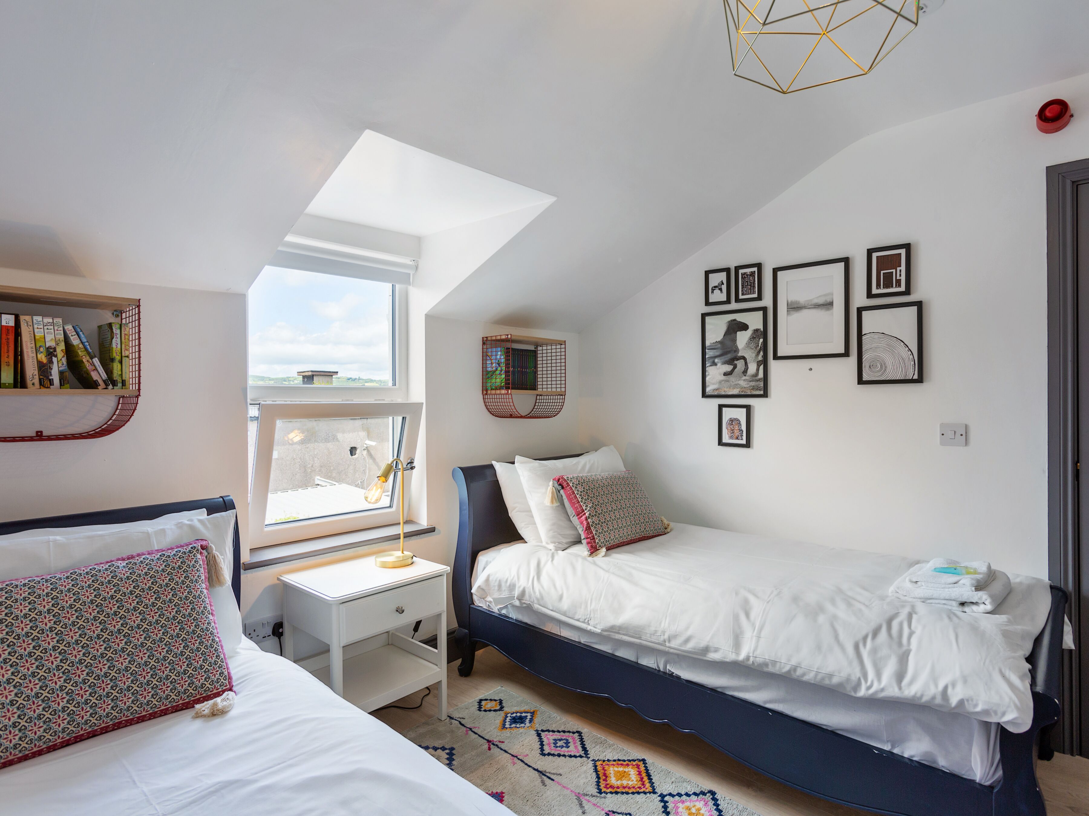 A bedroom with a skylight, a desk, a bed, and a wall with framed pictures.