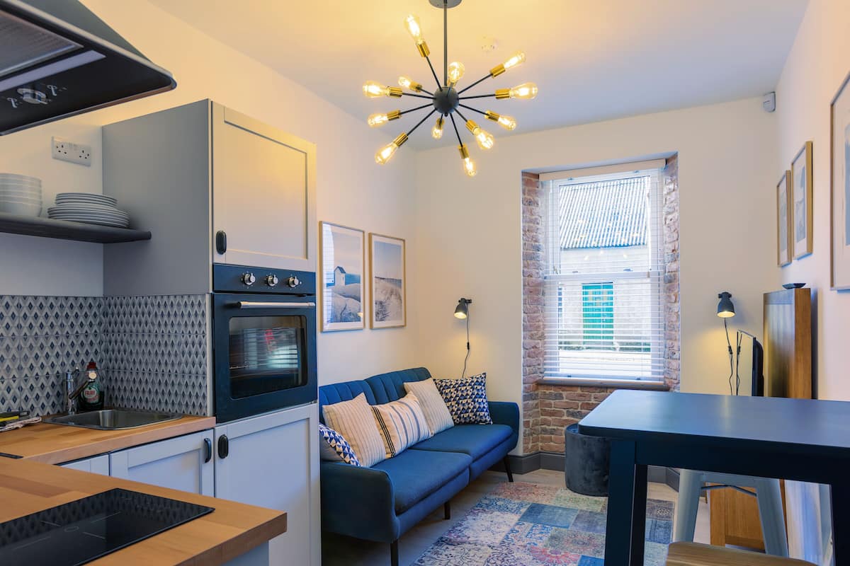 A modern kitchen with a built-in oven, a dining area with a table and chairs, a blue sofa, and a patterned rug.