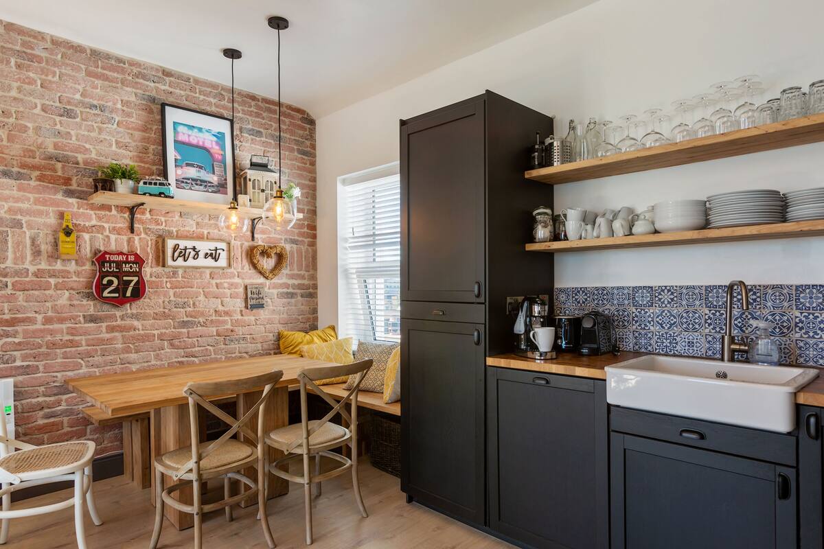 A kitchen with a brick wall, wooden dining table, and chairs. There are shelves with dishes and a coffee machine.