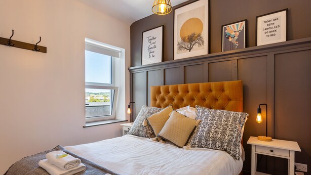 A neatly made bed with pillows and a folded towel, a bedside table with a lamp, and a window with a view. - Macs Townhouse (Dungarvan)