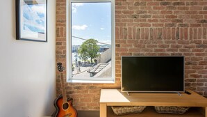 A room with a brick wall, a window overlooking a cityscape, a guitar, and a TV on a wooden stand. - Macs Townhouse (Dungarvan)