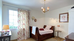 Un dormitorio con una cama de madera, un candelabro, una mesita de noche con lámpara, una ventana con cortinas florales y un cuadro en la pared.