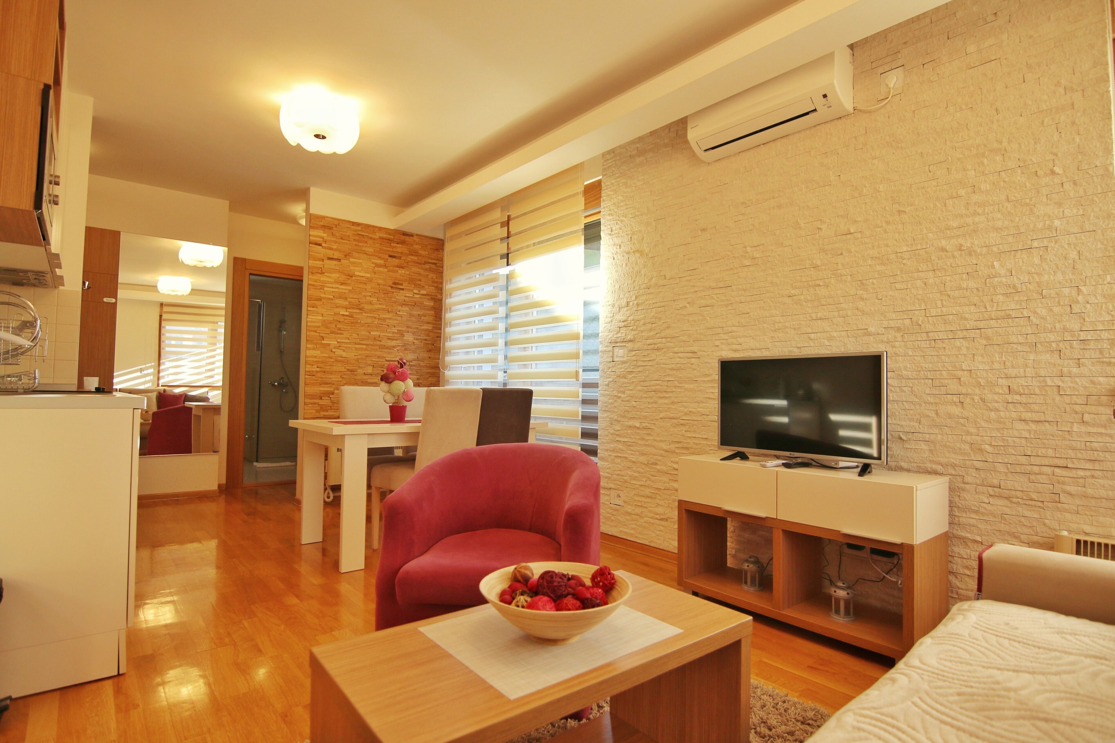 A modern living room with a wooden floor, a red armchair, a TV on a wooden stand, and a dining area with a table and chairs.