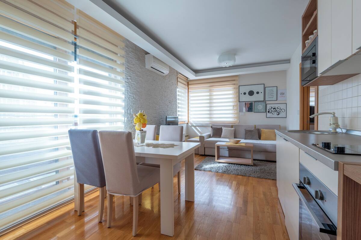 Cozinha moderna com área de jantar, piso de madeira e janelas amplas com persianas.