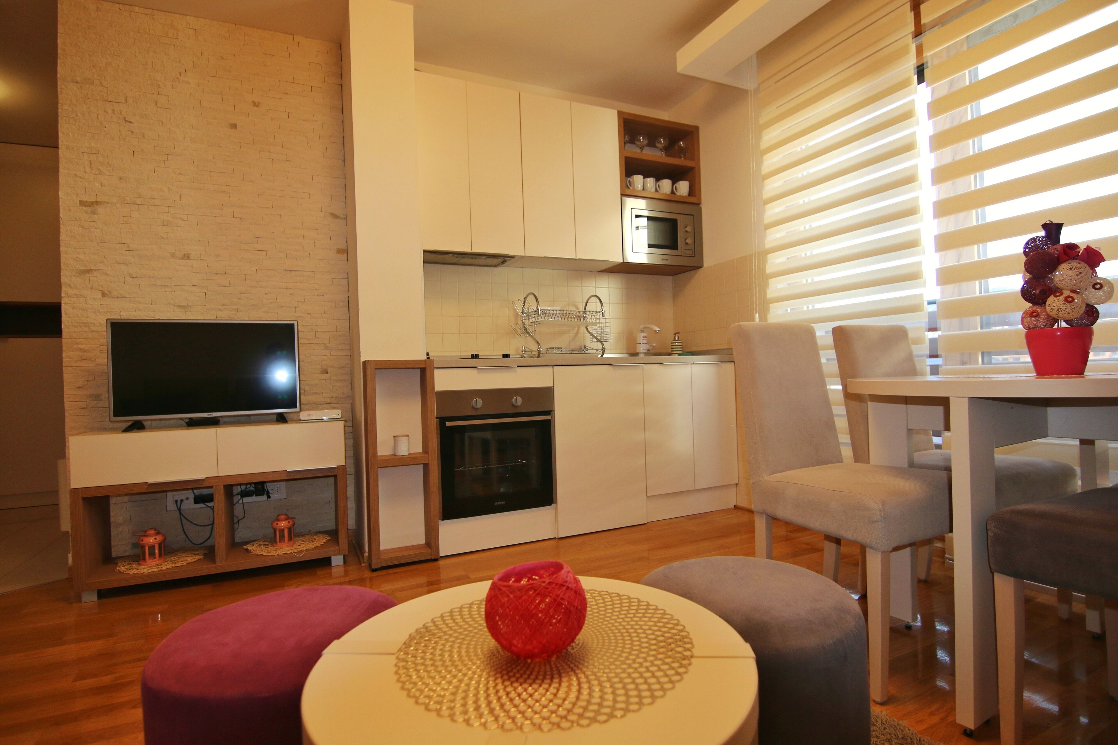 Une cuisine moderne avec des meubles blancs, un four à micro-ondes et un évier. On y trouve un coin repas avec une table et des chaises, ainsi qu’un téléviseur posé sur un meuble.
