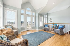 Il s’agit d’une pièce spacieuse avec un plafond haut, de grandes fenêtres et une vue sur l’extérieur. On y trouve des chaises en osier et un tapis bleu sur le sol.