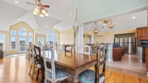 A spacious dining area with a wooden table and chairs, a kitchen with stainless steel appliances, and large windows allowing natural light. - Corolla Odyssey: Dog-Friendly, Elevator, Hot Tub, Private Pool (Corolla)