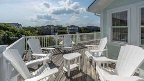 A wooden deck with white Adirondack chairs and a small table, overlooking a residential area with houses and greenery. - Corolla Odyssey: Dog-Friendly, Elevator, Hot Tub, Private Pool (Corolla)