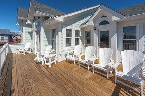Une terrasse en bois avec des fauteuils Adirondack blancs, un espace couvert avec une porte et une fenêtre équipée de stores.
