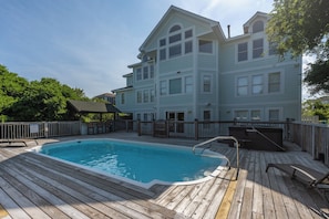 A swimming pool with a wooden deck, surrounded by a wooden fence and a two-story building with multiple windows. - Corolla Odyssey: Dog-Friendly, Elevator, Hot Tub, Private Pool (Corolla)