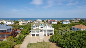 A large, two-story house with a prominent front porch and multiple balconies, surrounded by lush greenery and a view of the ocean. - Corolla Odyssey: Dog-Friendly, Elevator, Hot Tub, Private Pool (Corolla)
