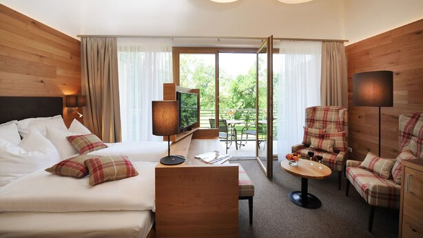 A hotel room with a bed, a TV, a seating area with a table, and a balcony with a view of greenery. - Landgasthof Karolinenhöhe (Lichtenfels)